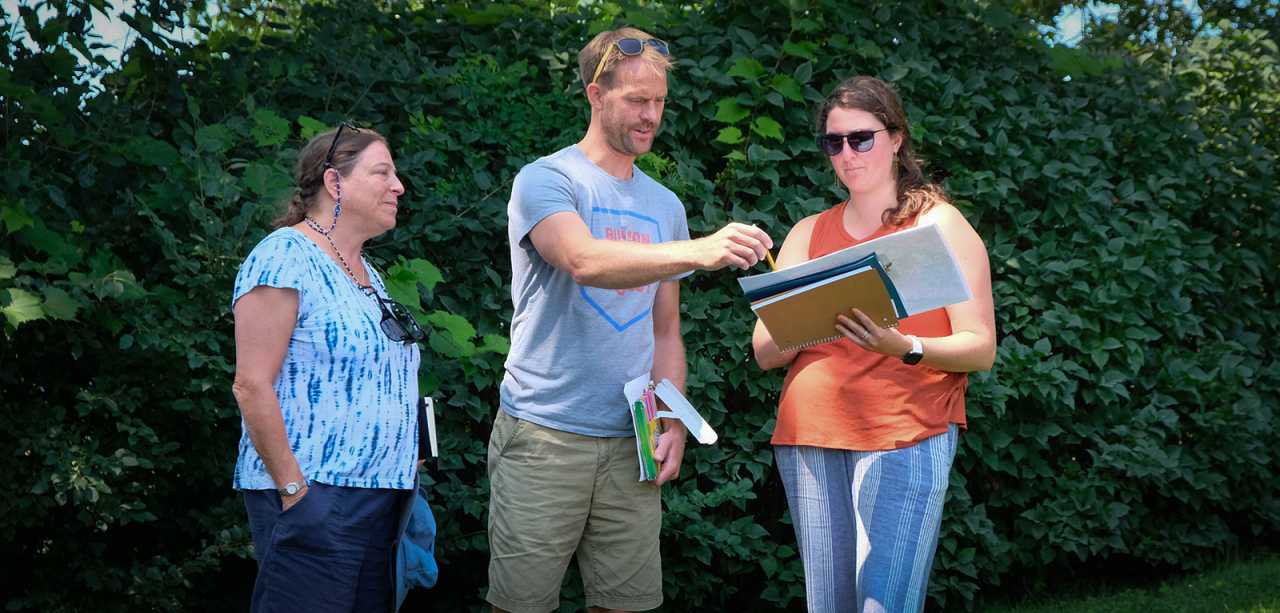Three people stand outside against a green hedge in summertime, looking at and gesturing at a clipboard collaboratively