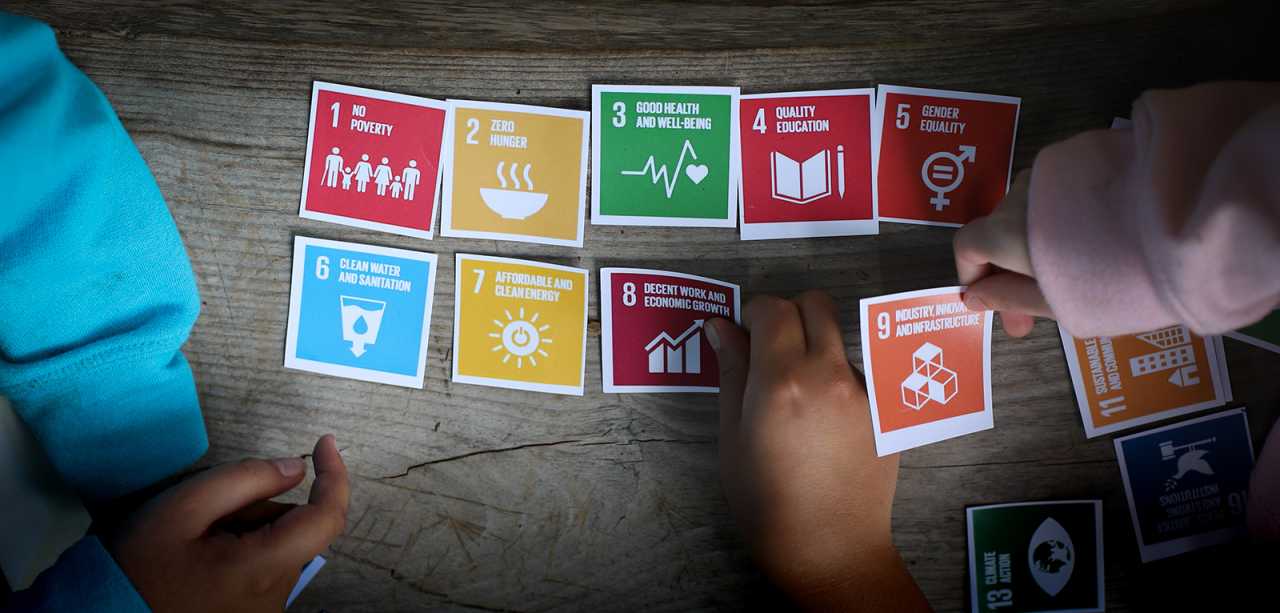 Colorful cards with icons representing the sustainable development goals are spread out on a table, with three sets of hands touching the cards