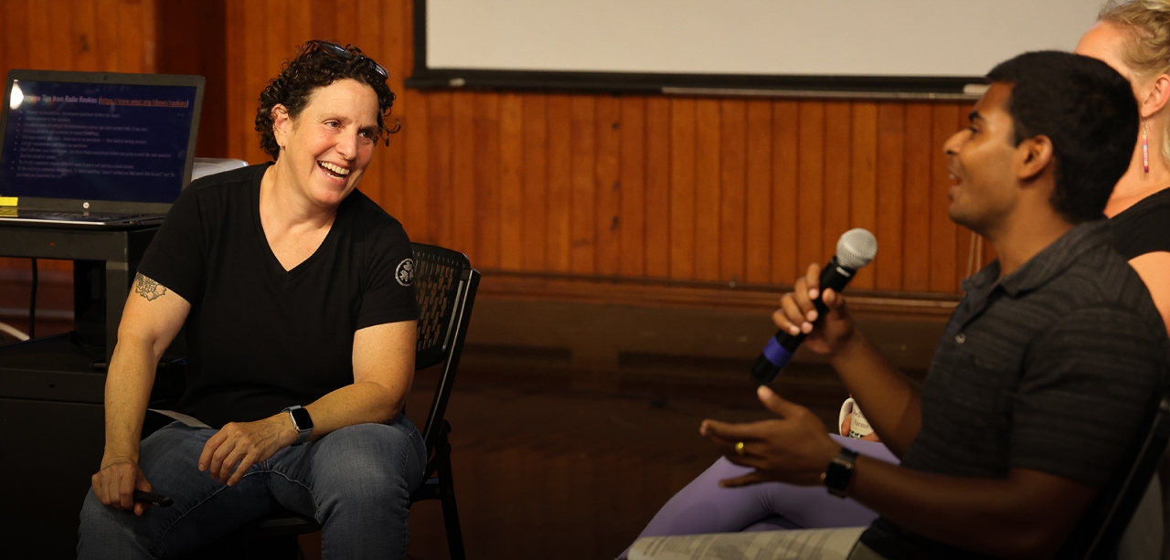 Two people smile while in conversation, one holding a microphone, in a wood paneled room