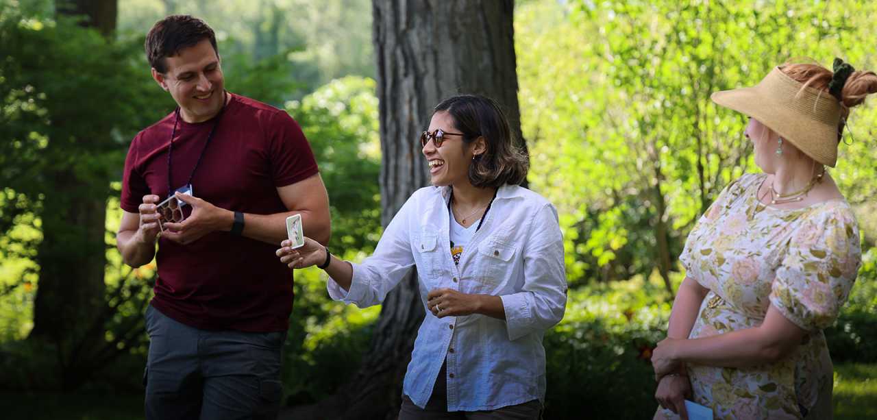 Three people stand in a row outdoors shaded by leafy green trees laughing while displaying illustrated cards