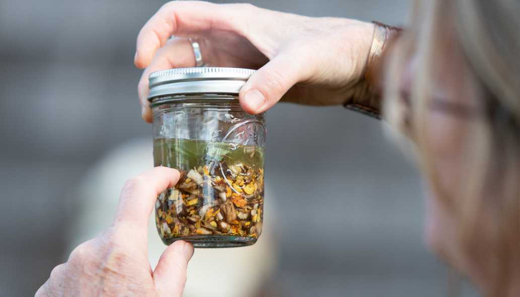 A person holds up a lidded glass jar, full of herbal ingredients and steeped in a clear liquid.