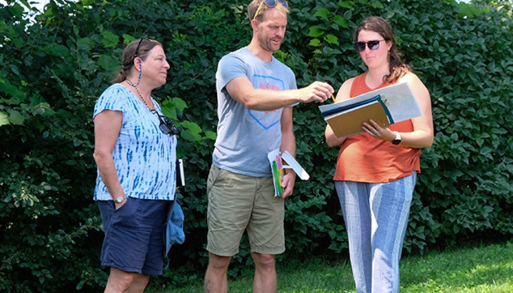 Three people stand looking at a clipboard outside, standing in front of a green hedge