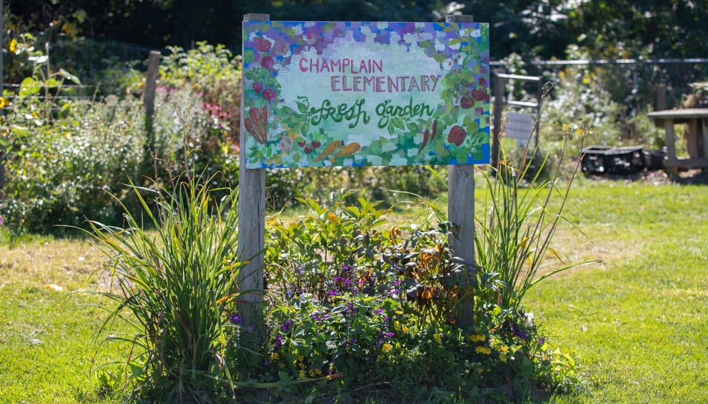 A hand painted sign with "Champlain Elementary Fresh Garden". A garden is in the background.