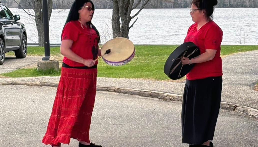 Two women wearing red playing hand drums in front of a body of water.