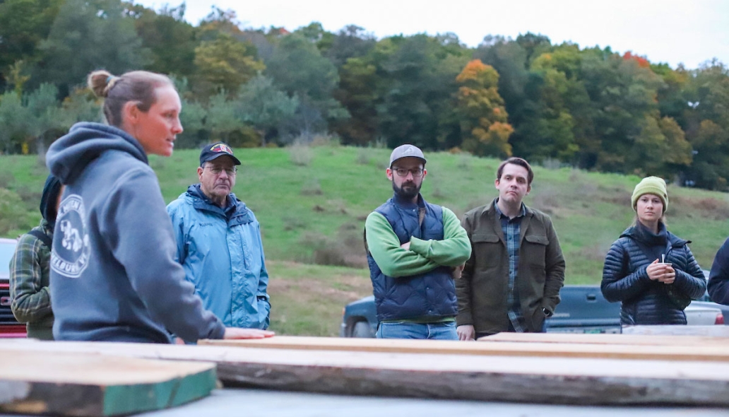 People gather around a collection of lumber outdoors, one person speaks to the group.