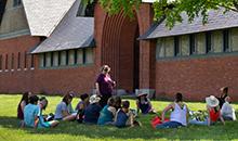 A dozen educators sit in shaded grass in front of the historic brick Coach Barn at Shelburne Farms