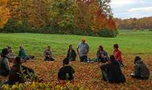 A dozen educators sit in a circle on an expansive green lawn with autumn foliage in distance