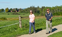 a woman in man in sunglasses, short sleeves, and long pants walk side by side on a gravel trail beside pastures