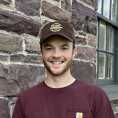 Smiling young man with short beard, t-shirt, and baseball cap against a wall of redstone. Shown shoulders and up