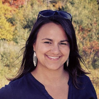 Woman with dark hair and sunglasses on head smiles at the camera in front of a forest background.