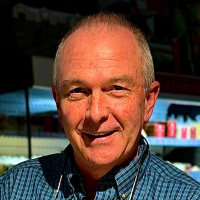 Man with short gray hair in a blue shirt smiles at the camera, a refrigerated market display behind him.