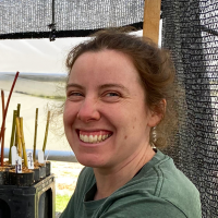 Woman with brown hair, pulled back, in a green shirt smiles at camera with plant cuttings in the background.