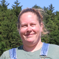 Woman with blonde hair, pulled back, wearing denim overalls and green t-shirt smiles at the camera in front of green trees and a blue sky.