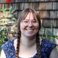 Woman with long bangs, round wire frame glasses, and light brown hair in two braids smiles at the camera in front of a natural backdrop with tall greenery.