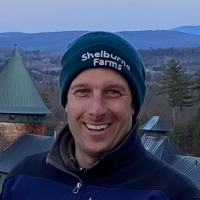 Man in "Shelburne Farms" beanie smiles at the camera with views of the property in the background.