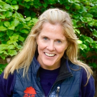 Woman with long blonde hair smiles at the camera in front of a leafy background.