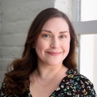 Woman with long brown hair smiles at camera in front of a white wall.