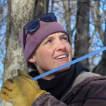 Woman in a light purple beanie and yellow gloves smiling in a forested area.