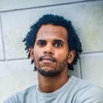 Person with dark hair curly hair and facial hair looks into the camera in front of a white stone wall.