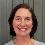 Woman with a brown bob smiles at the camera in front of a background of muted green paneling.