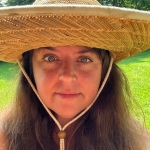 Woman in a wide-brimmed straw hat smiles at the camera in a grassy setting.
