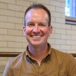 Man with short blonde hair smiles wearing a tan shirt.