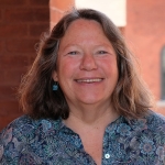Woman with shoulder length brown hair and blue eyes smiles at camera in an outdoor setting.