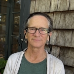 woman with graying hair pulled back, wearing glasses with a strap, and a white cardigan over a green tshirt.