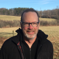 Man with short light-brown hair, dark frame glasses and a short salt and pepper beard smiles at the camera with a field of cold weather grass and trees behind him.