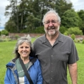 A middle-aged couple standing together outside. The man, with glasses and gray beard, has his arm around the woman's shoulder. 