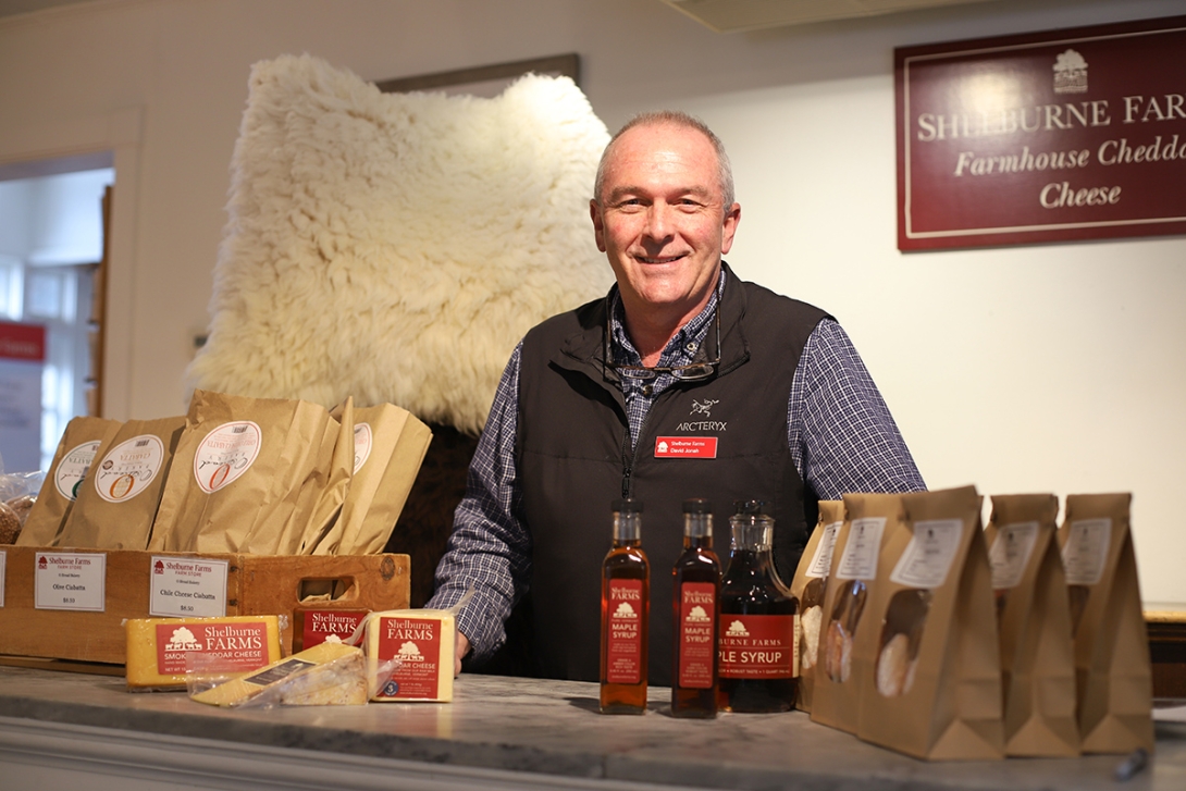 Man in fleece vest behind counter that has cheese, bread, and syrup on it. Sheepskins behind him