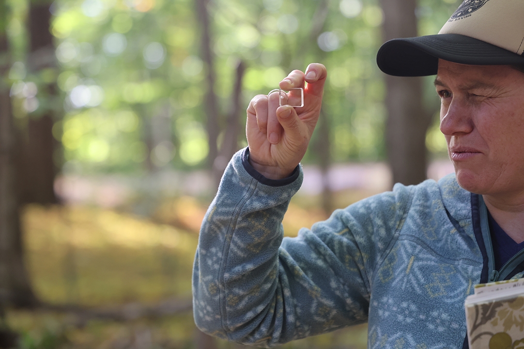 A person holds a small prism and squints to gaze through it while standing in a golden autumn forest